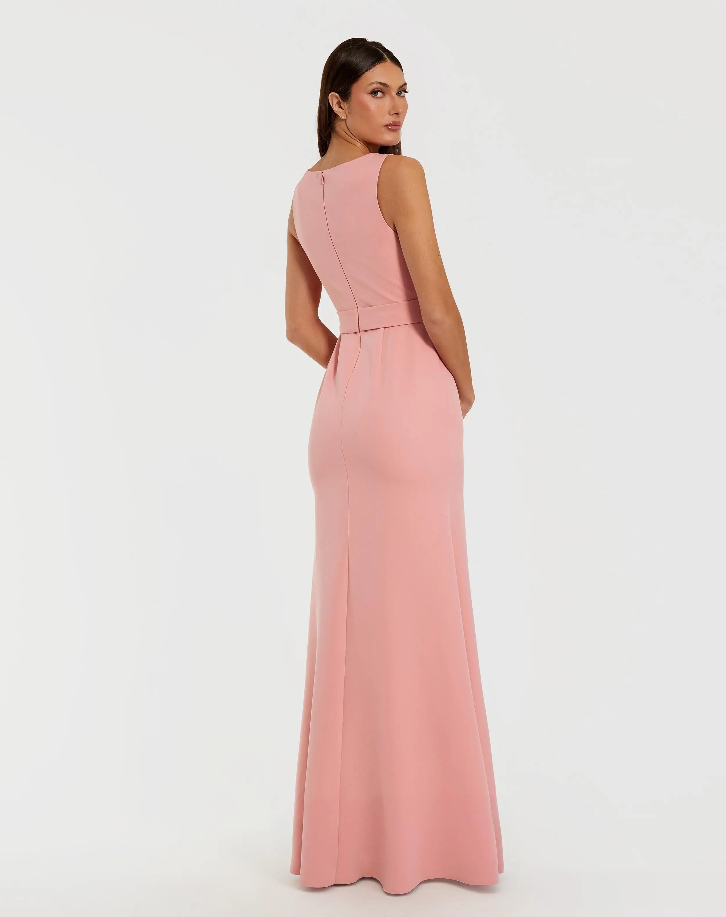 A woman is seen from behind, glancing over her left shoulder while wearing the Mac Duggal 50097 Long Formal Dress—a sleeveless, floor-length crepe gown in a light pink pastel shade—set against a plain white background.
